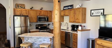 A modern kitchen with wooden cabinets, a stainless steel refrigerator, a microwave, a stove, and a sink. There are two wooden stools and a sign on the wall.