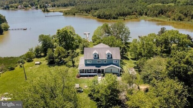 Une grande maison dotée d’un porche avant imposant, entourée d’un lac et d’une forêt dense.