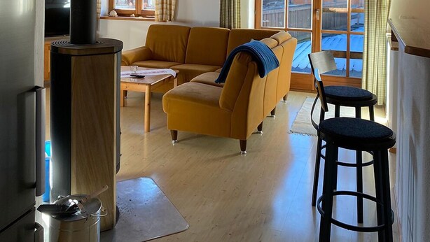 Een gezellige woonkamer met een houten plafond, een bank, een tafel, en een keuken met een fornuis.