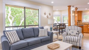 A living room with a gray sofa, striped armchair, and a wooden coffee table. There is a dining area with a table and chairs, and a kitchen with wooden cabinets and a window.