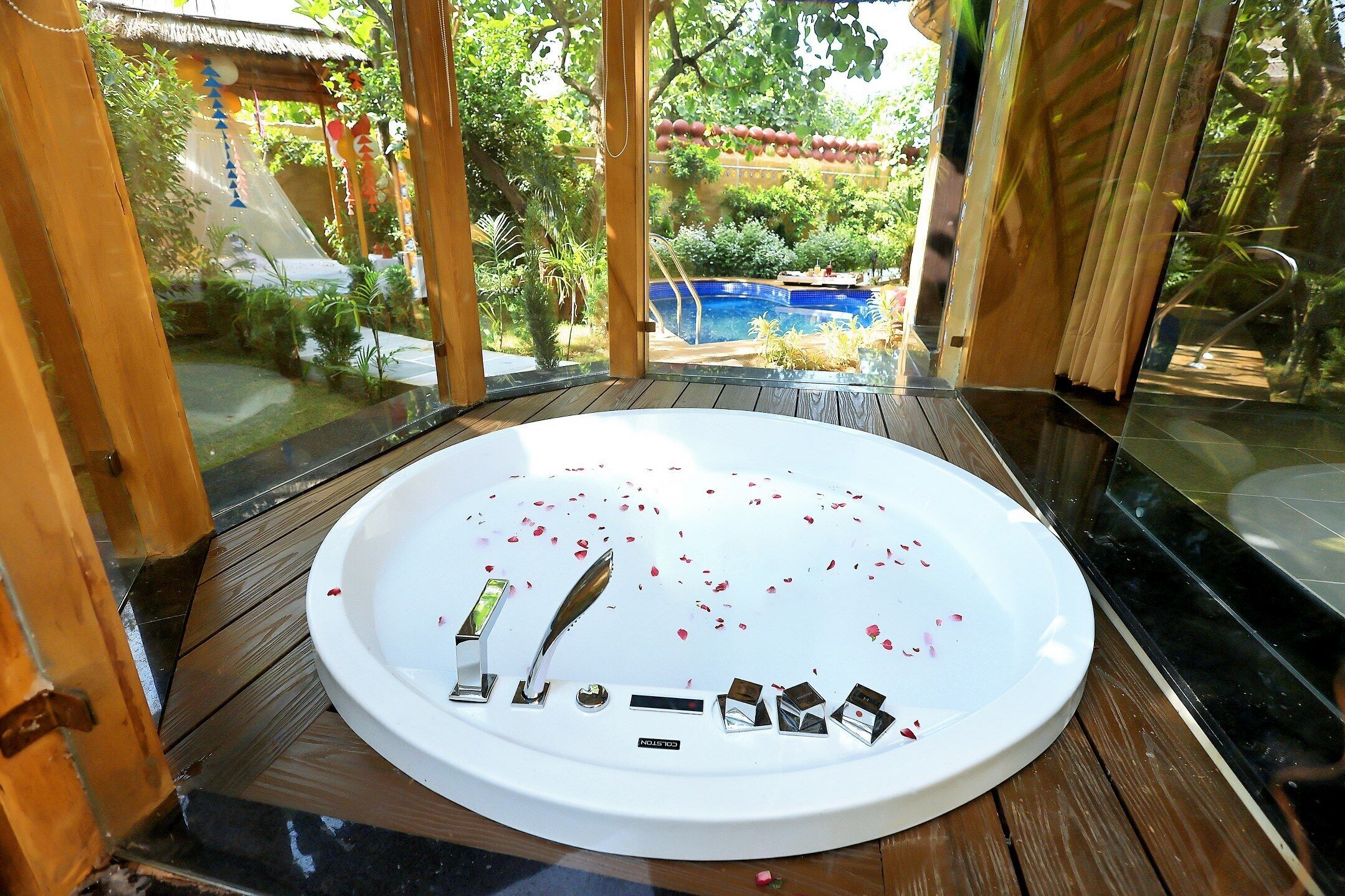 A circular hot tub with rose petals and a remote control on the edge, surrounded by a wooden deck and a glass enclosure.