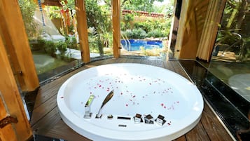 A circular hot tub with rose petals and a remote control on the edge, surrounded by a wooden deck and a glass enclosure.