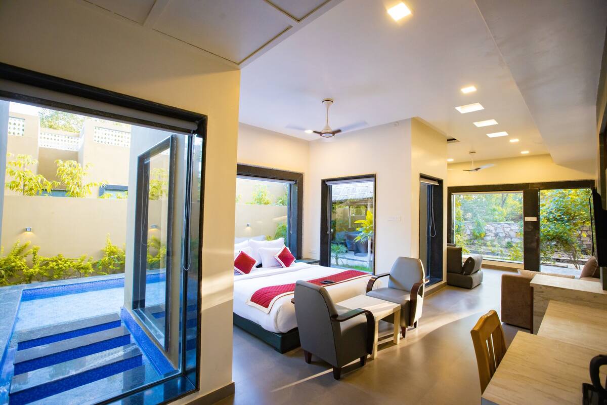 A modern hotel room with a large bed, a seating area, and a balcony with a view of greenery.