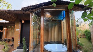 Ein mit Glas umgebenes Whirlpool mit Holzterrasse, umgeben von einem Garten und einem Gebäude mit Strohdach.