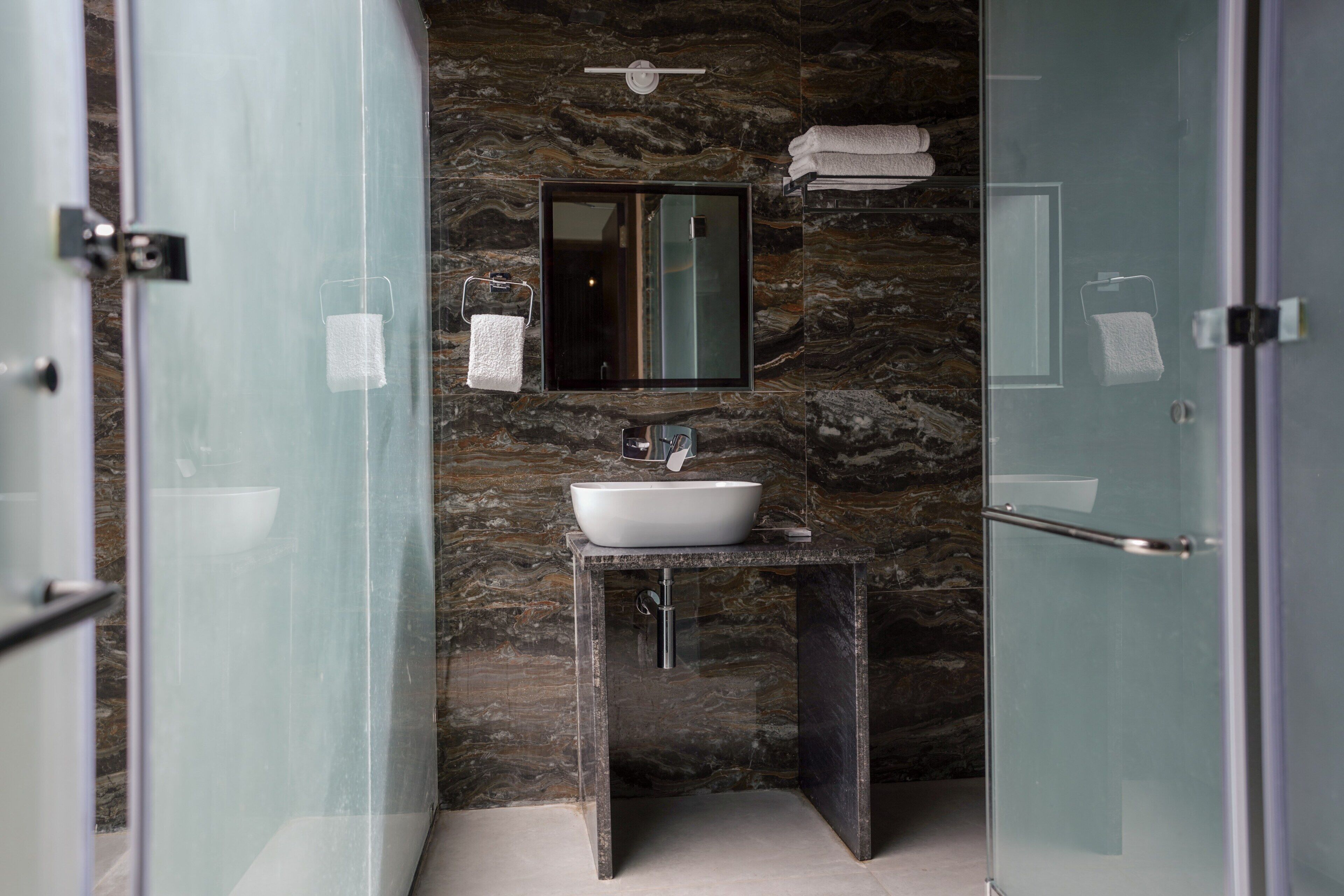 Un baño con cabina de ducha de vidrio, pared de mármol, lavamanos con espejo y un perchero para toallas.