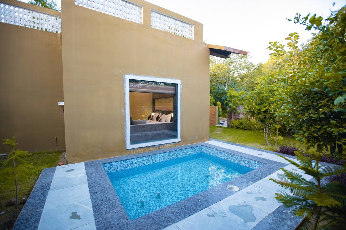 A swimming pool with a glass enclosure, surrounded by a garden with various plants and a building with a beige exterior.