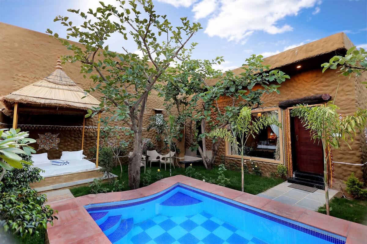 A pool area with a checkered blue tile pattern, surrounded by a garden with a thatched-roof structure and a wooden door.