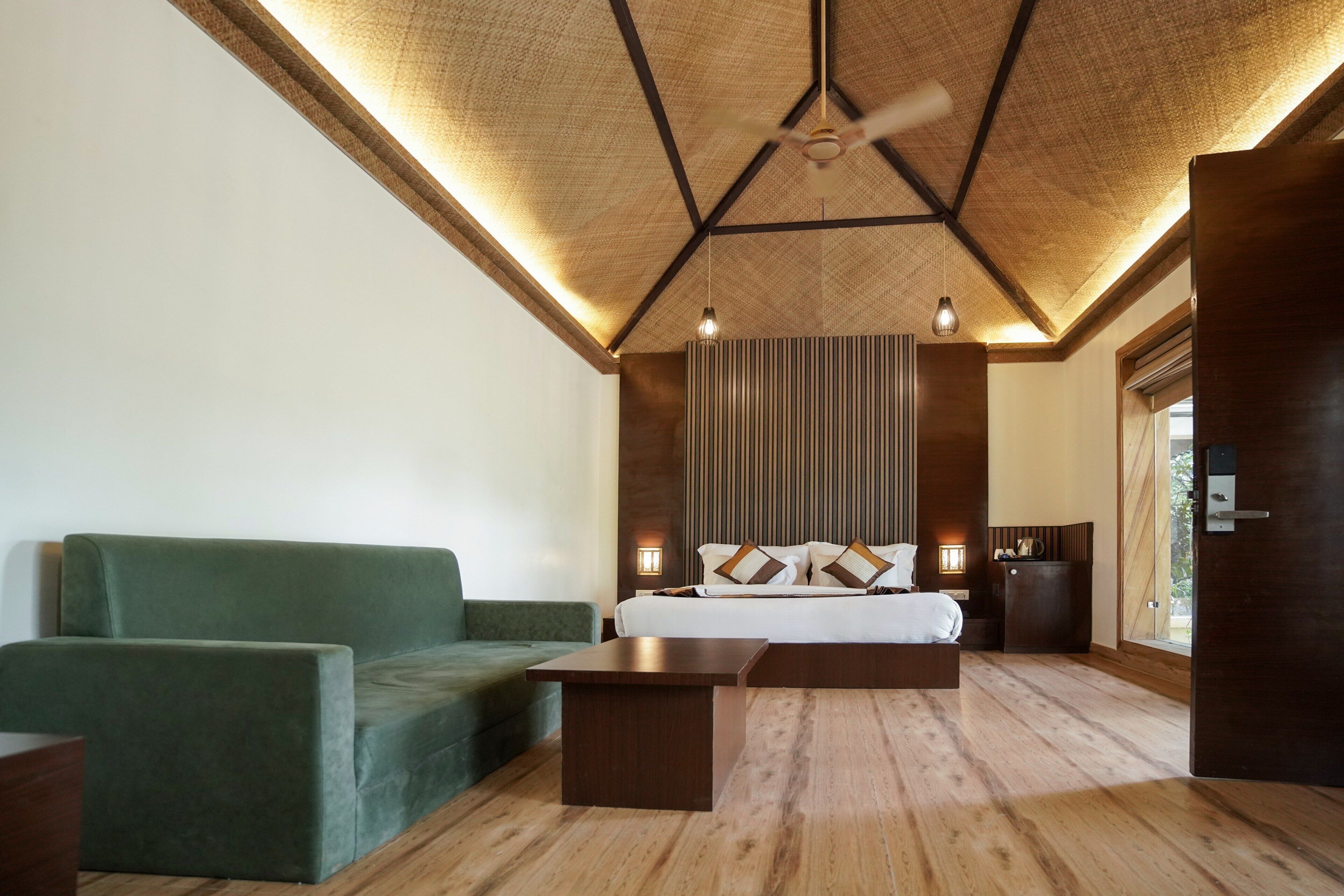 A modern hotel room with a wooden ceiling, a green sofa, a wooden coffee table, a bed with white linens, and a wall-mounted light fixture.
