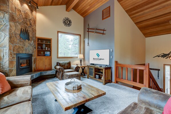 A cozy living room with a stone fireplace, leather armchairs, a wooden coffee table, and a large window offering a view of trees. - COYO18 - Beautiful, Private Hot Tub, Gas Fireplace (Sunriver)