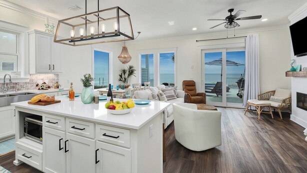 A modern kitchen with white cabinetry, a central island with fruit bowl, and a dining area with a view of the ocean. - On Tide Time - Beachfront | Private Pool | SAT-SAT (Panama City Beach)
