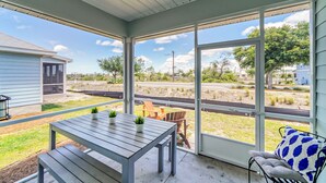A covered patio with a table and chairs, large windows offering a view of a road and greenery. - Port St. Joe Home | Heated Pool | Dog Friendly | Sleeps 10 (Port St Joe)