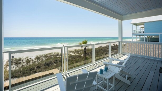Una terraza de madera con sillas y una mesa blancas, con vista a una playa y al océano.