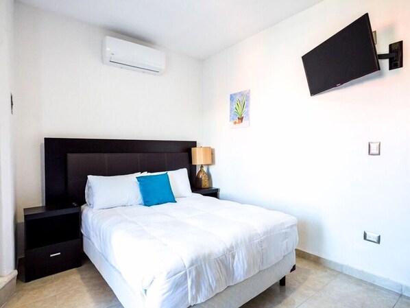 A bedroom with a bed, a nightstand, a lamp, an air conditioner, a TV mounted on the wall, and a small plant. - Loft Oasis (Playa del Carmen)