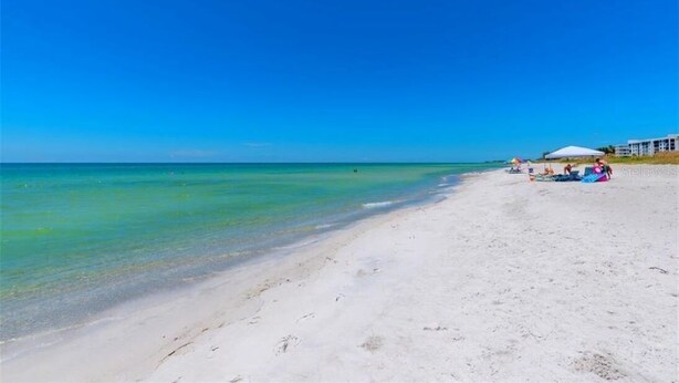 Een strand met helder turquoise water, wit zand en enkele personen en parasols.