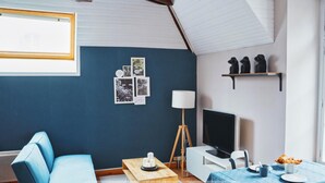 A room with a blue wall, a wooden ceiling, a blue sofa, a table with a lamp, a TV, and a shelf with figurines. - Le Woody - Proche Gare - Télétravail (Laval)