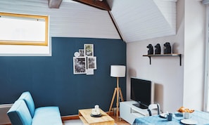 Ein Zimmer mit blauer Wand, Holzboden, einem blauen Sofa, einem Tisch mit Lampe, einem Fernseher und einem Regal mit Figuren.