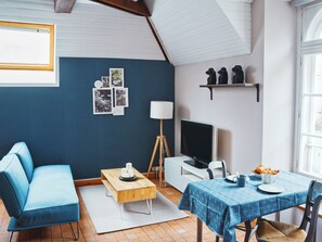 Ein Zimmer mit einem blauen Sofa, einem Holztisch, einem Fernseher auf einem Schrank, einem Esstisch mit blauer Tischdecke und einer Wand mit gerahmten Fotos.