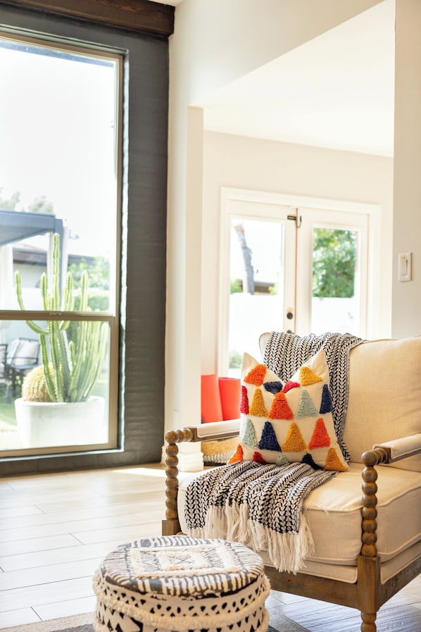A room with a large window, a chair with a throw blanket, a patterned ottoman, and a cactus plant.