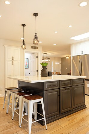 Una cocina moderna con una isla central, taburetes de bar, iluminación colgante y un refrigerador de acero inoxidable.