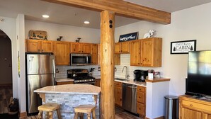 Una cocina moderna con armarios de madera, un refrigerador de acero inoxidable, un microondas, una estufa y un lavamanos. Hay dos taburetes y un pilar de madera.
