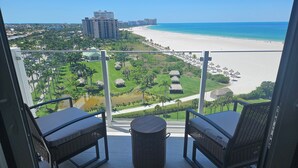 A balcony with two wicker chairs and a small table, overlooking a beach with a clear view of the ocean and a resort area with thatched-roof huts. - Sunny Oceanfront Condominium, Short Walk to the Beach (Marco Island)
