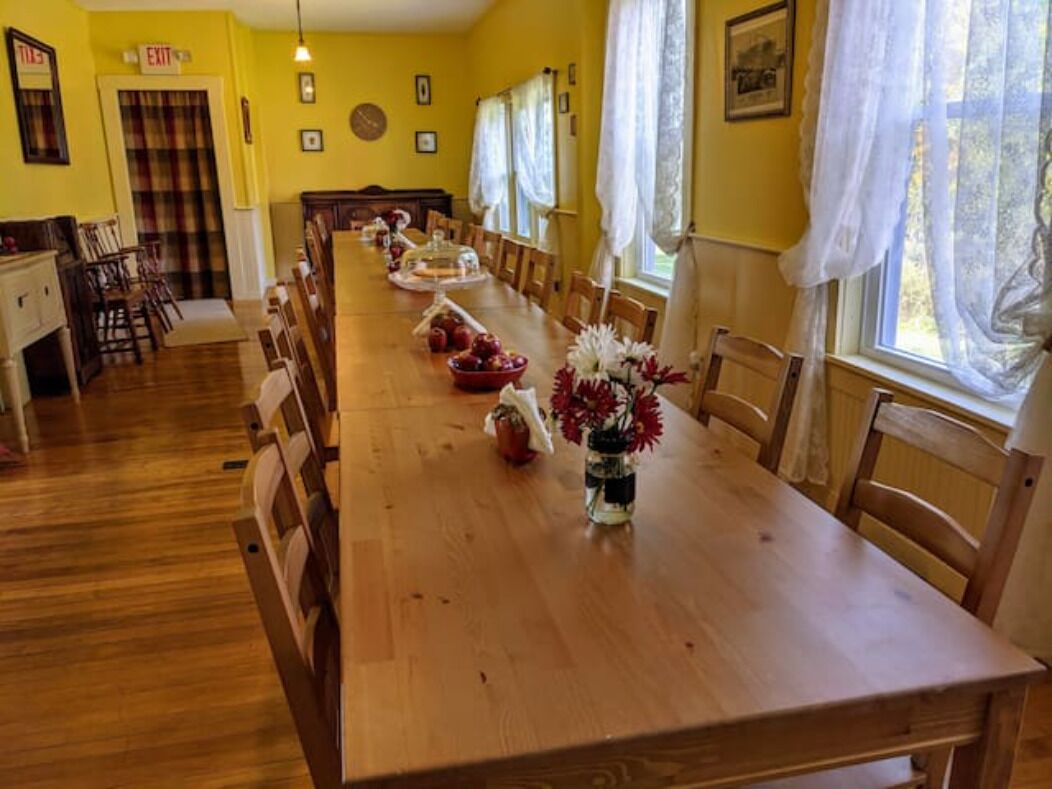 A long wooden dining table with chairs, a vase of flowers, and a buffet setup in a room with yellow walls and large windows.