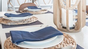 Ein Essbereich mit einem langen Tisch, gedeckt für zwei Personen, mit gewebten Tischsets, Tellern und blauen Servietten. Über dem Tisch hängt eine hölzerne Laterne als Leuchte.