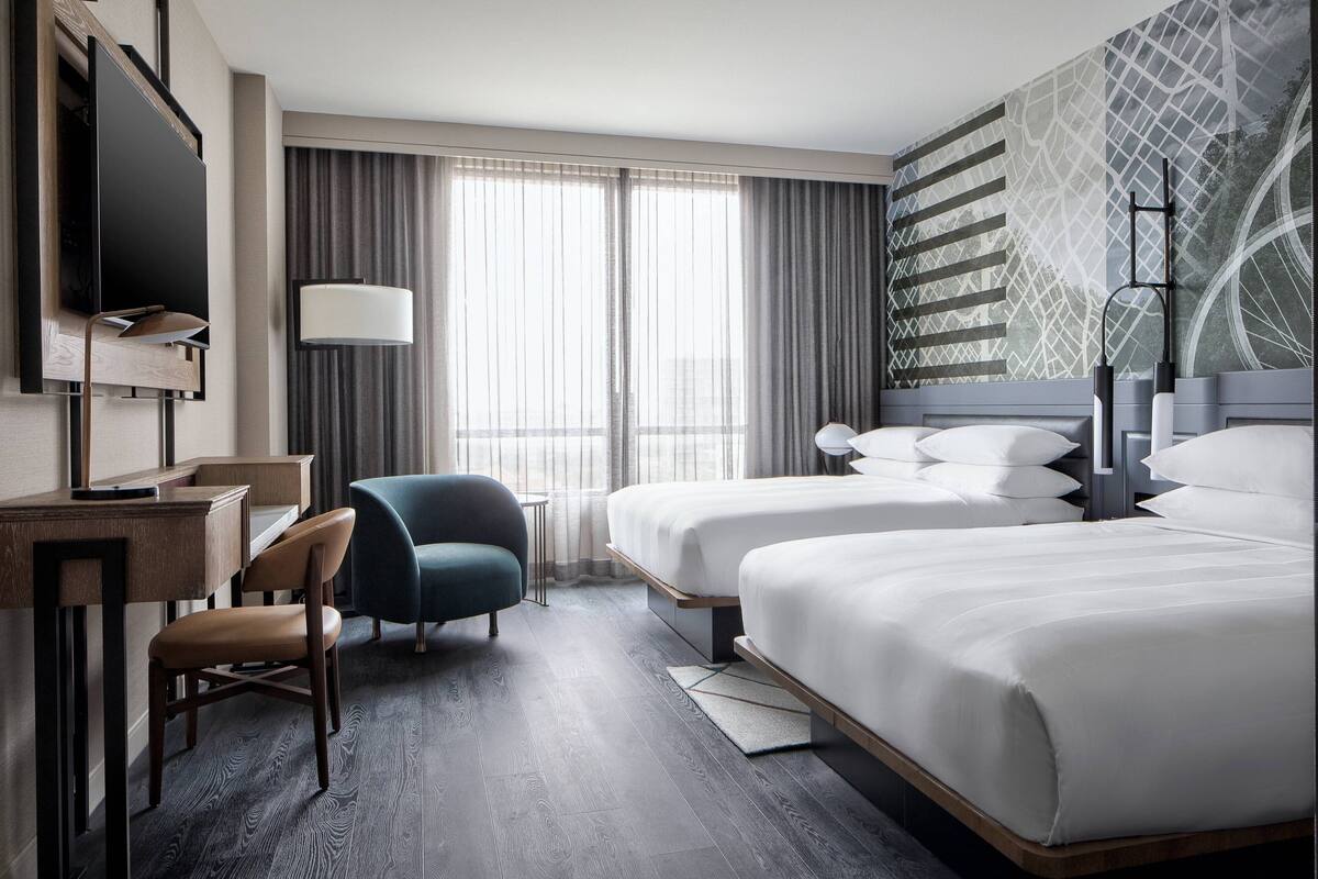 Ein Hotelzimmer mit Schreibtisch, zwei Betten, einem Sessel und einem großen Fenster mit Vorhängen.