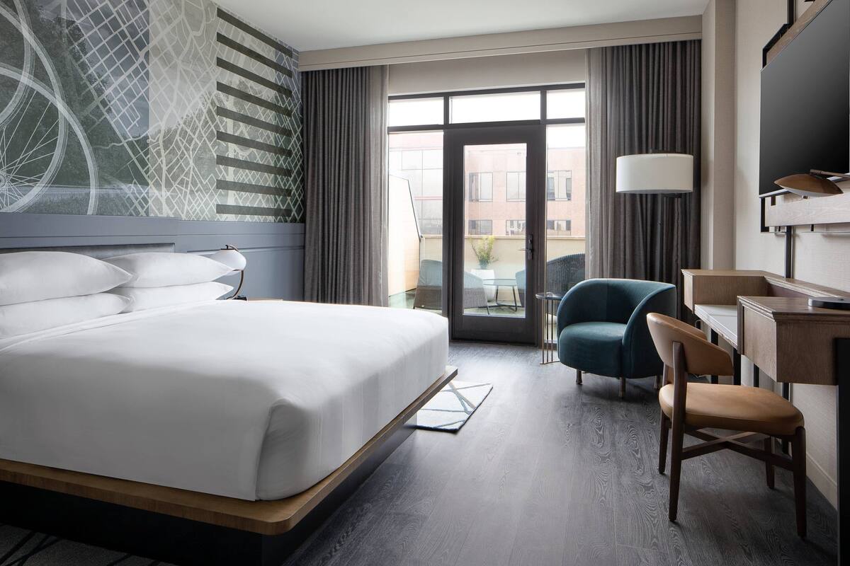 Ein Hotelzimmer mit einem großen Bett, einem Schreibtisch, einem Sessel und Blick ins Freie.
