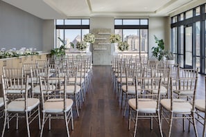 A spacious indoor venue with rows of bamboo chairs arranged for an event, large windows offering a city view, and a decorative floral arrangement at the front.