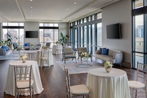 A spacious hotel room with large windows, a flat-screen TV, multiple seating areas, and a dining table.