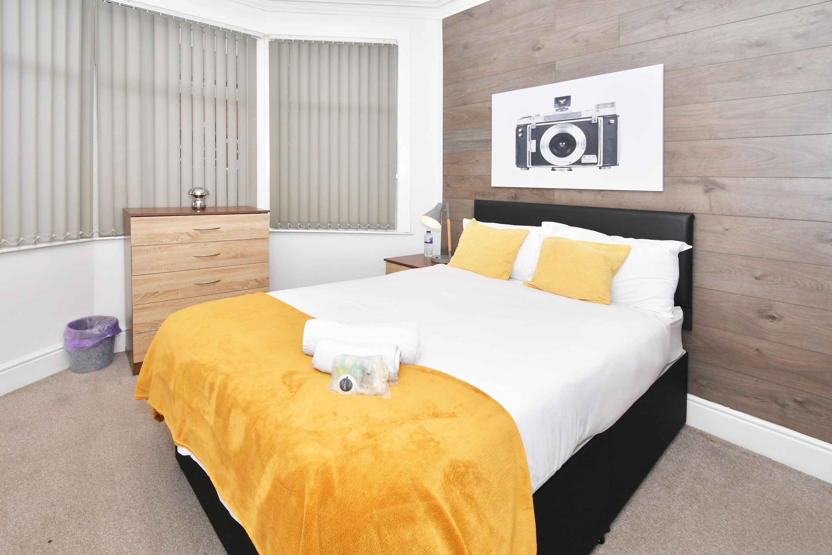 A bedroom with a bed, a wooden dresser, a nightstand, a camera poster, and a window with blinds.