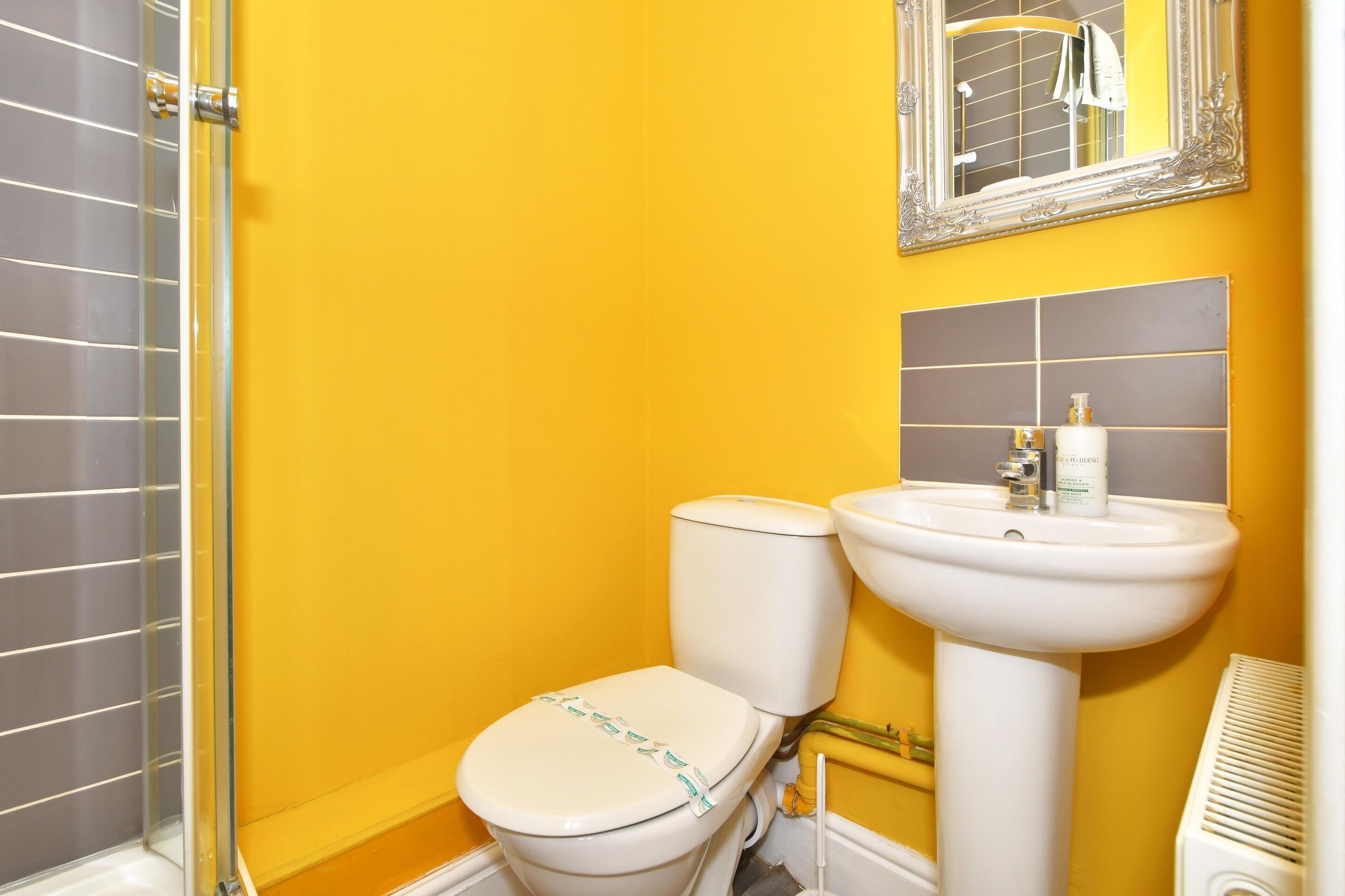 Et badeværelse med gul væg, et hvidt toilet, en hvid vask og et spejl over vasken.