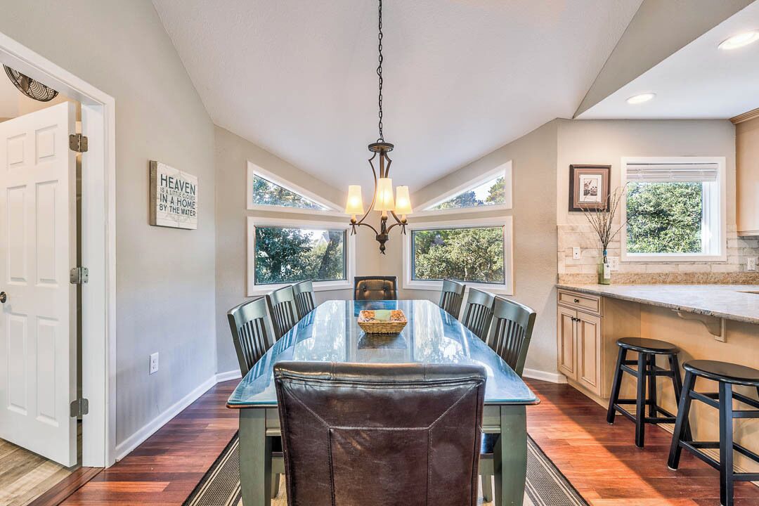 Une salle à manger avec une grande table, des chaises et un lustre. On peut voir des fenêtres et un coin cuisine.