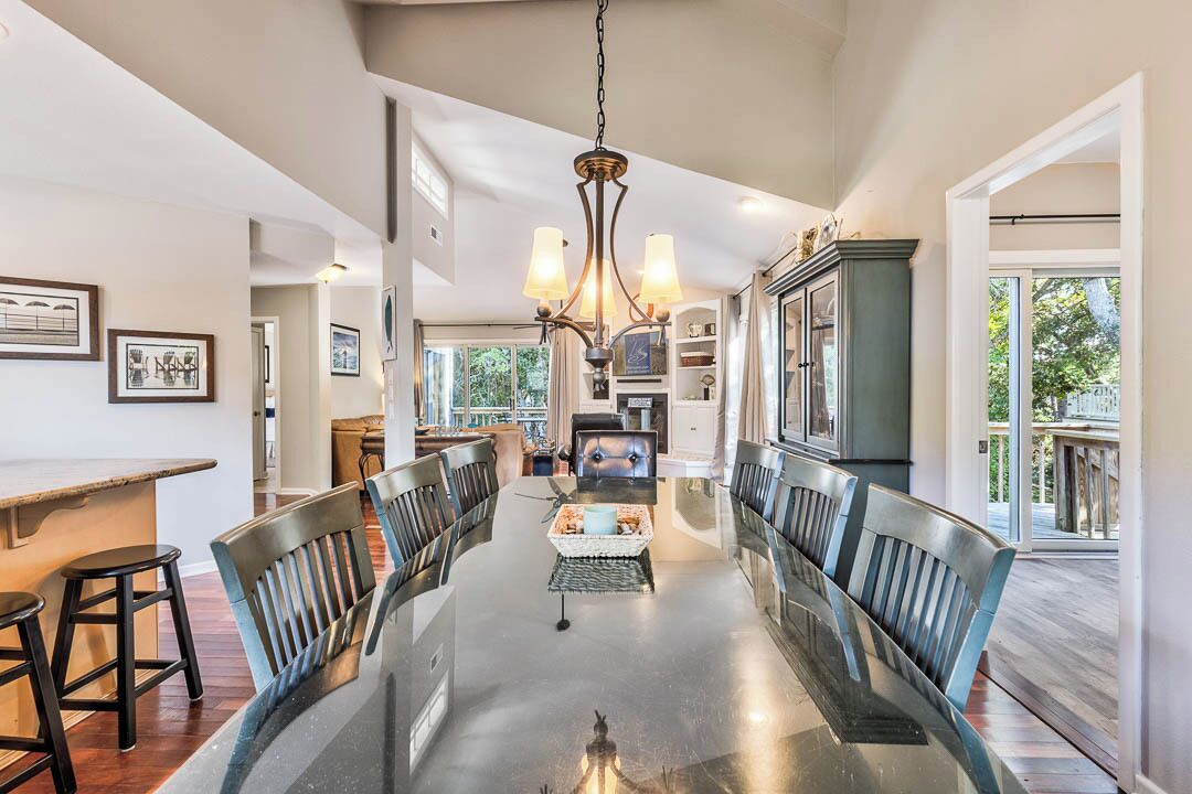 A spacious dining area with a long table, chairs, and a chandelier. There is a kitchen in the background with a window overlooking greenery.