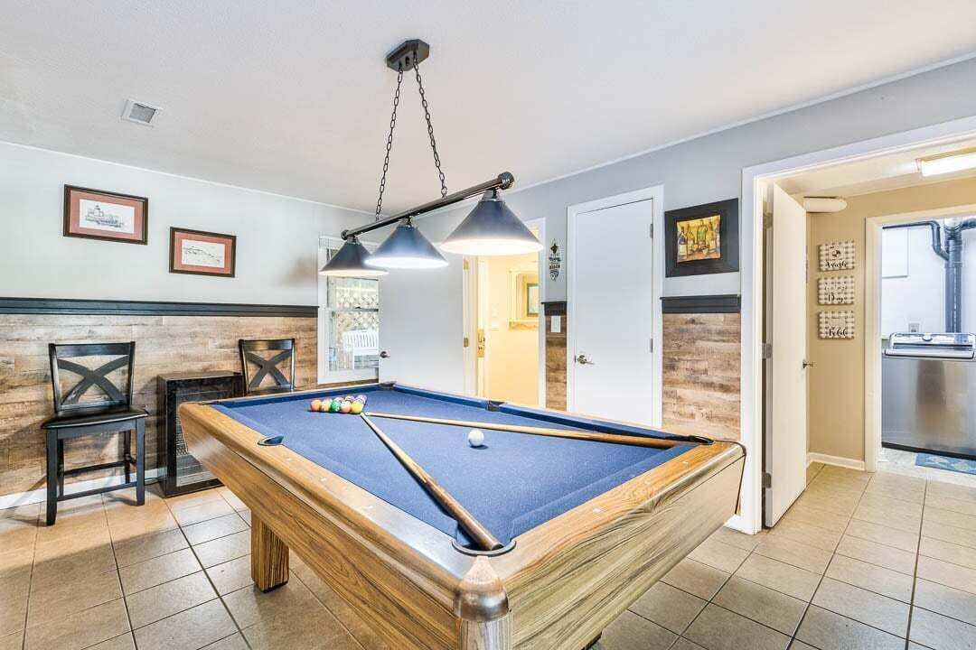 A pool table in a room with a wooden floor, a ceiling light fixture, and framed pictures on the wall.