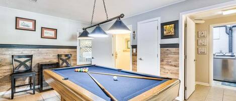 A pool table in a room with a wooden floor, a ceiling light fixture, and framed pictures on the wall.