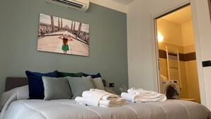 Hotellihuone, jossa on sänky, tyynyjä, taulu ja avoimesta ovesta näkyvä kylpyhuone.