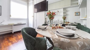Une salle à manger moderne avec une table ronde dressée pour quatre personnes, des chaises à revêtement vert, un téléviseur à écran plat fixé au mur et une fenêtre laissant entrer la lumière naturelle.