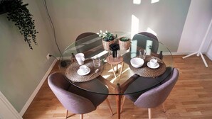 Une table de salle à manger avec un plateau en verre, accompagnée de deux chaises violettes, entourée de sets de table en fibres tressées, de tasses blanches et d’une petite plante en pot.