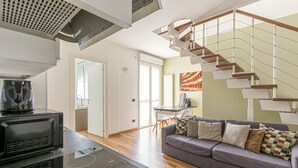 A modern kitchen with a built-in stove, a dining area with a table and chairs, a gray sofa with patterned cushions, and a unique staircase design. - Candiani Building - Elegant 2-level Apartment with 3 bedroms (Milano)