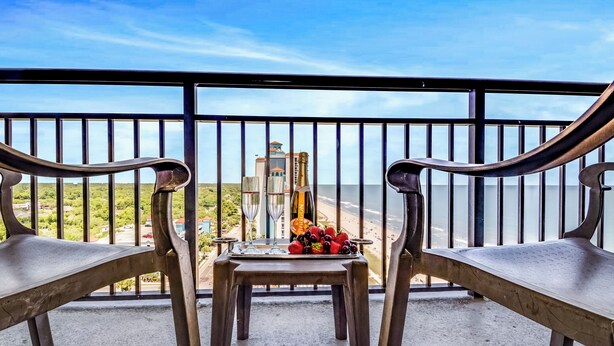 A balcony with two metal chairs, a small table with a tray of fruit and two glasses, and a view of the beach and buildings in the distance. - Relaxation Awaits/1Br Suite Dogs OK! (Myrtle Beach)