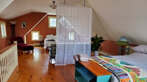 Un dormitorio con suelo de madera, una cama con colcha de colores, una silla, una mesa, una planta en maceta y una escalera.