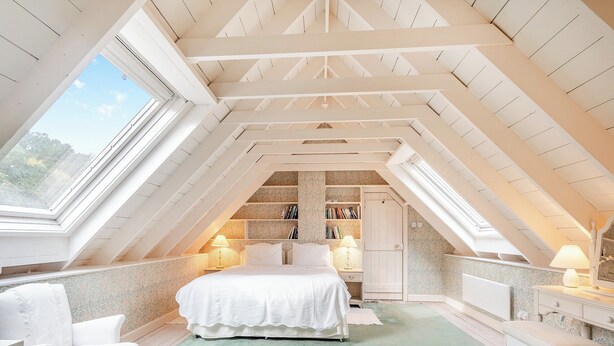 Une chambre mansardée chaleureuse, avec un lit, deux chaises, un banc et une bibliothèque.