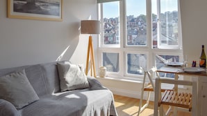 A cozy living room with a sofa, a lamp, and a view of a cityscape through large windows. - 1 bedroom accommodation in Swanage (Swanage)