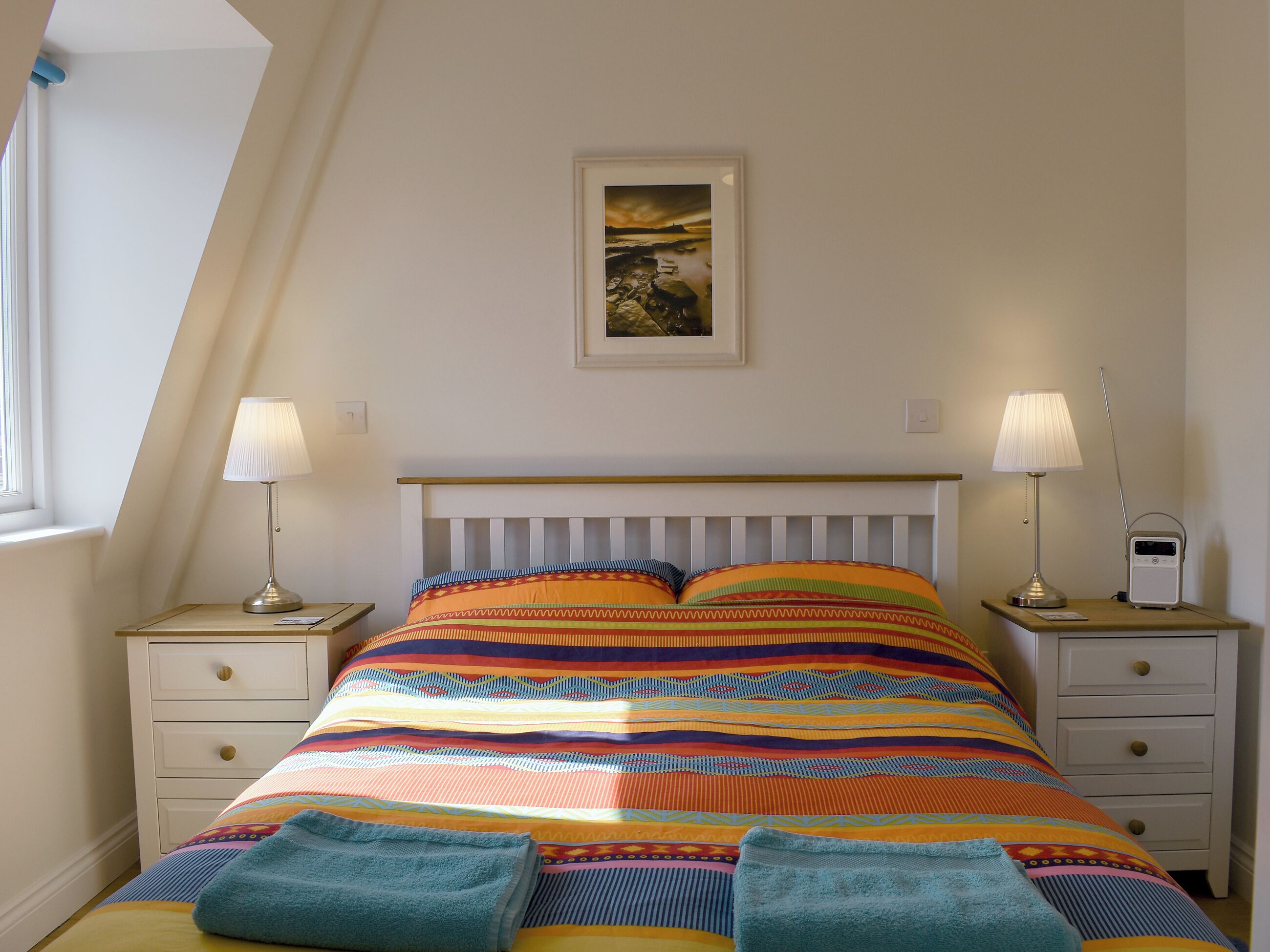 Een slaapkamer met een bed, twee nachtkastjes, een lamp, een radio en een schilderij aan de muur.