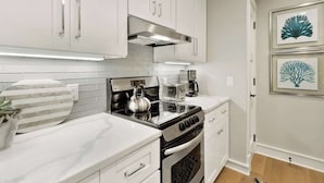Una cocina moderna con armarios blancos, una campana de acero inoxidable, una estufa a gas, una tetera y una cafetera. Hay obras de arte enmarcadas en la pared y una planta en maceta sobre la encimera.