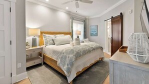 Un dormitorio con cama, mesitas de noche, ventilador de techo, ventana con persianas, puerta de madera y televisor montado en la pared.