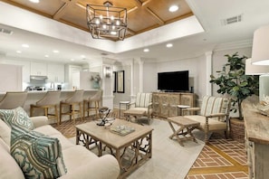 A spacious living area with a sofa, chairs, a coffee table, a TV, and a kitchen in the background.