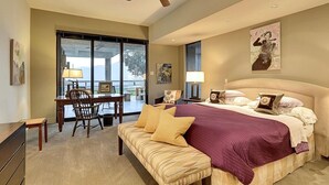 A bedroom with a large bed, a desk, and a chair. There is a ceiling fan, a window, and artwork on the wall.